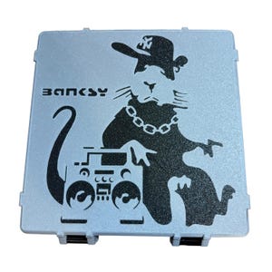 May include: A light blue, rectangular case featuring a black graphic of a rat wearing a hat and chain, leaning on a boombox. The word "BANKSY" is printed above the rat. The case has a textured surface.