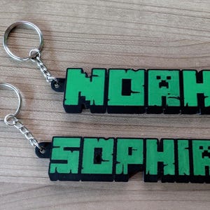 Puede incluir: Dos llaveros verdes y negros con los nombres "Noah" y "Sophia" en una fuente inspirada en Minecraft. Cada llavero tiene un anillo de llave plateado.