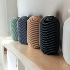 Könnte beinhalten: Fünf dekorative Vasen in einer Reihe, jede mit einer einzigartigen Farbe und Design. Die Vasen sind zylindrisch mit gerippter Textur und die Farben umfassen Grün, Blau, Beige und Dunkeltürkis. Sie stehen auf einer hellen Holzoberfläche.