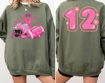 May include: An olive green sweatshirt with a pink football helmet, football, and awareness ribbon graphic on the front. The back of the sweatshirt features the number 12 in pink with floral accents. The word "Navy" is printed on the bottom left.