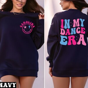 May include: A navy blue sweatshirt featuring a front graphic with a pink smiley face, a star, a lightning bolt, and the name Carolina. The back displays the text In My Dance Era in large, wavy, pink and light blue letters with small star accents.
