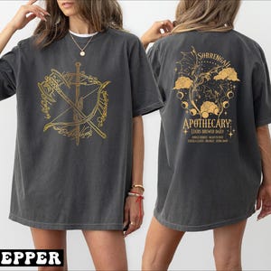 May include: Dark grey t-shirt with a gold graphic on the front featuring a sword, axe, and bow within a circular design. The back of the shirt has a gold apothecary design with text. The word "PEPPER" is at the bottom of the image.