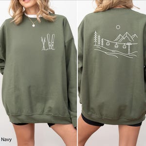 May include: An olive green crewneck sweatshirt featuring a white line art graphic of skis and poles on the left chest. The back displays a white line art illustration of a mountain landscape with pine trees, a ski lift, and a sun.