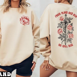 May include: Sand-colored sweatshirt with a red rose graphic on the front. The back features a sword design with roses and the text "FROM BLOOD AND ASH WE WILL RISE" in red. A casual, comfortable top.
