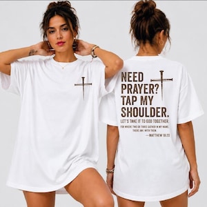 A woman wearing a white t-shirt with a cross and text printed on it, posing with her hands on her shoulders.