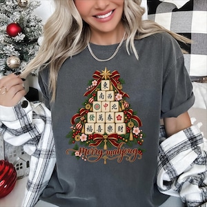 Puede incluir: Una camiseta gris carbón con un gráfico de un árbol de Navidad formado por fichas de mahjong. El árbol está decorado con cintas rojas, borlas doradas y flores. El texto "Merry mahjong" aparece en letra roja debajo del árbol.