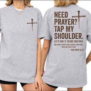 two gray t-shirts with a Bible verse printed on the back. The text on the back of the shirts reads "NEED PRAYER? TAP MY SHoulder. LET'S TAKE IT TO GOD TOGETHER. FOR WHERE TWO THIRTS GATHER IN MY NAME, THERE IS WITH THEM - Matthew 18:20".