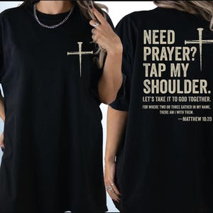two black t-shirts with white text and a cross design. The text on the back of the shirts reads "NEED PRAYER? TAP MY SHoulder. LET'S TAKE IT TO GOD TOGETHER. FOR WHERE TWO OR THREE GATHER IN MY NAME, THERE AM I WITH THEM. -MATTHEW 18:20".