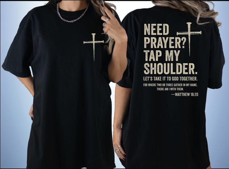 two black t-shirts with white text and a cross design. The text on the back of the shirts reads "NEED PRAYER? TAP MY SHoulder. LET'S TAKE IT TO GOD TOGETHER. FOR WHERE TWO OR THREE GATHER IN MY NAME, THERE AM I WITH THEM. -MATTHEW 18:20".