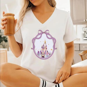 A woman wearing a white t-shirt with a castle graphic and a purple ribbon design, holding a drink in her hand.