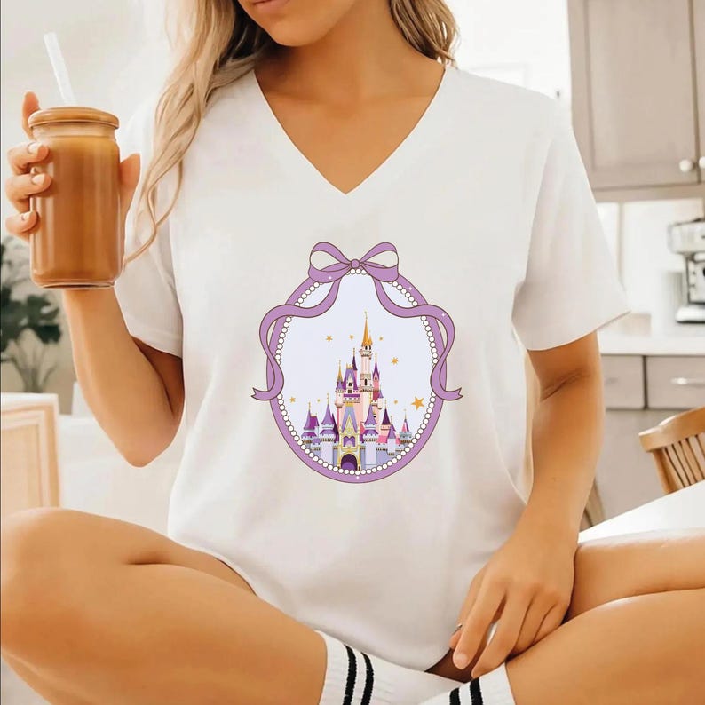 A woman wearing a white t-shirt with a castle graphic and a purple ribbon design, holding a drink in her hand.
