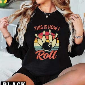May include: Black sweatshirt featuring a bowling-themed graphic. The design includes bowling pins, a bowling ball, and the text "THIS IS HOW I ROLL" in a retro color scheme. The word "BLACK" is at the bottom of the image.