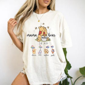 Puede incluir: Una camiseta de manga corta color blanco roto con un gráfico de Winnie the Pooh con un tarro de miel, el texto mama bear, Est. 2025, y cuatro personajes más pequeños sosteniendo globos con los nombres oliver, mia, sophia y jack impresos debajo.