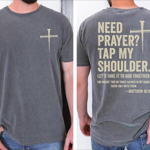 a man wearing a gray t-shirt with a Bible verse printed on the back. The text on the t-shirt reads "NEED PRAYER? TAP MY SHoulder. LET'S TAKE IT TO GOD TOGETHER. FOR WHERE TWO OR THREE GATHER IN MY NAME, THERE I AM WITH THEM. -MATTHEW 18:20".