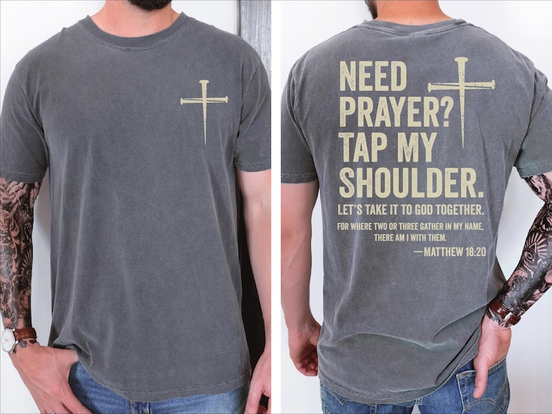 a man wearing a gray t-shirt with a Bible verse printed on the back. The text on the t-shirt reads "NEED PRAYER? TAP MY SHoulder. LET'S TAKE IT TO GOD TOGETHER. FOR WHERE TWO OR THREE GATHER IN MY NAME, THERE I AM WITH THEM. -MATTHEW 18:20".