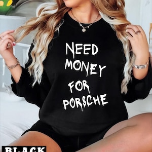 May include: A black long-sleeve sweatshirt featuring the white text NEED MONEY FOR PORSCHE in a stylized, dripping font. The garment has a crew neckline and is worn with matching black shorts, creating a coordinated set with a bold graphic design.