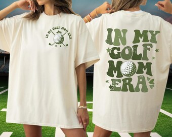May include: Cream-colored t-shirt with the text "IN MY GOLF MOM ERA" in green. The front features a golf ball and crossed clubs. The back of the shirt repeats the phrase with stars and a golf ball graphic. A casual, comfortable tee.