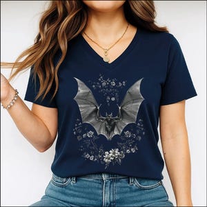 May include: A navy blue V-neck t-shirt featuring a central graphic of a grey bat with outstretched wings, surrounded by a detailed arrangement of white and grey floral elements. The design is printed on the front of the short-sleeved garment.