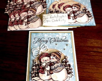 May include: A set of handmade greeting cards featuring a printed illustration of two snowmen wearing hats and scarves, with the text Merry Christmas. The cards include white twine bows, small embellishments, and a note labelled Tan Pearls/crystals.