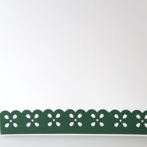 May include: A white greeting card with a dark green scalloped border featuring a floral design. The card is open, revealing a blank white interior, perfect for a personalized message. The border adds a decorative touch.