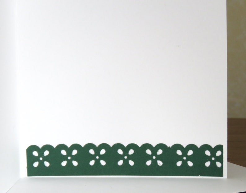 May include: A white greeting card with a dark green scalloped border featuring a floral design. The card is open, revealing a blank white interior, perfect for a personalized message. The border adds a decorative touch.