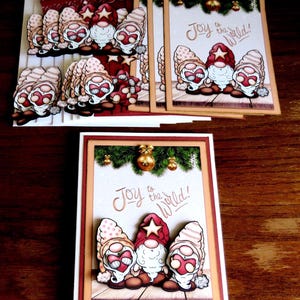 May include: Christmas-themed greeting cards featuring gnome illustrations. The cards have a white background with the phrase "Joy to the World!" and are decorated with festive elements like ornaments and greenery. The gnomes wear red and beige hats.
