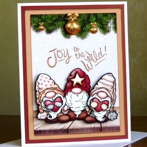 May include: A Christmas card featuring three cartoon gnomes with red hats and white beards. The card has the phrase "Joy to the World!" in gold lettering. The background includes a wooden floor, green pine branches, and gold ornaments.