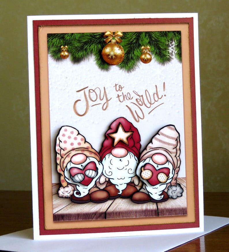May include: A Christmas card featuring three cartoon gnomes with red hats and white beards. The card has the phrase "Joy to the World!" in gold lettering. The background includes a wooden floor, green pine branches, and gold ornaments.