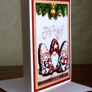 May include: A Christmas greeting card featuring three gnome figures with red hats and white beards. The card has the phrase "Joy to the World!" written on it, with a festive background of green garland and gold ornaments.