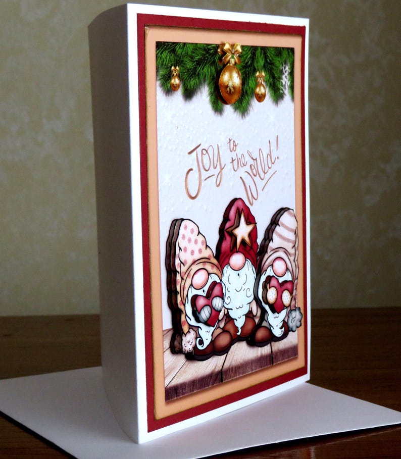 May include: A Christmas greeting card featuring three gnome figures with red hats and white beards. The card has the phrase "Joy to the World!" written on it, with a festive background of green garland and gold ornaments.