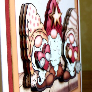 May include: A handmade greeting card featuring three whimsical gnomes. The gnomes have red, pink, and brown hats, holding red hearts. The card has a layered design with a wooden plank background and a cream-colored border.