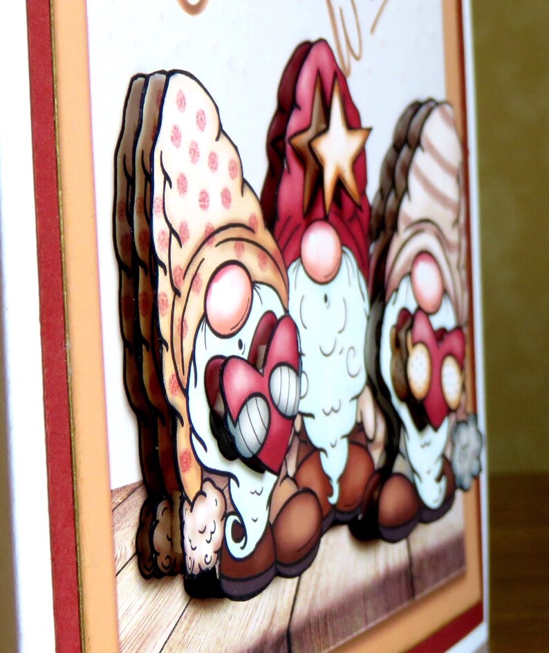 May include: A handmade greeting card featuring three whimsical gnomes. The gnomes have red, pink, and brown hats, holding red hearts. The card has a layered design with a wooden plank background and a cream-colored border.