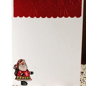 Op de afbeelding: Een witte wenskaart met een rode, in reli&euml;f aangebrachte rand aan de bovenzijde, voorzien van een geschulpte rand en een strikpatroon. In de linkerbenedenhoek staat een kleine, kleurrijke illustratie van de Kerstman in een rode jas.