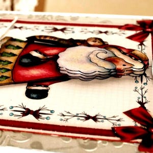 Op de afbeelding: Een handgemaakte wenskaart met een gelaagde, driedimensionale illustratie van de Kerstman in een rode jas met een witte baard. De kaart heeft een cr&egrave;mekleurige achtergrond, rode randen, decoratieve rode strikken en een label met Merry Christmas.