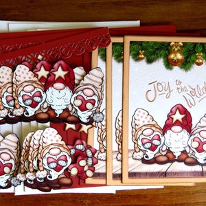 May include: Christmas greeting cards featuring gnome illustrations. The gnomes wear red hats with stars and have white beards. One card has the text "Joy to the World!" and a green border with gold ornaments. The cards are on a wooden surface.