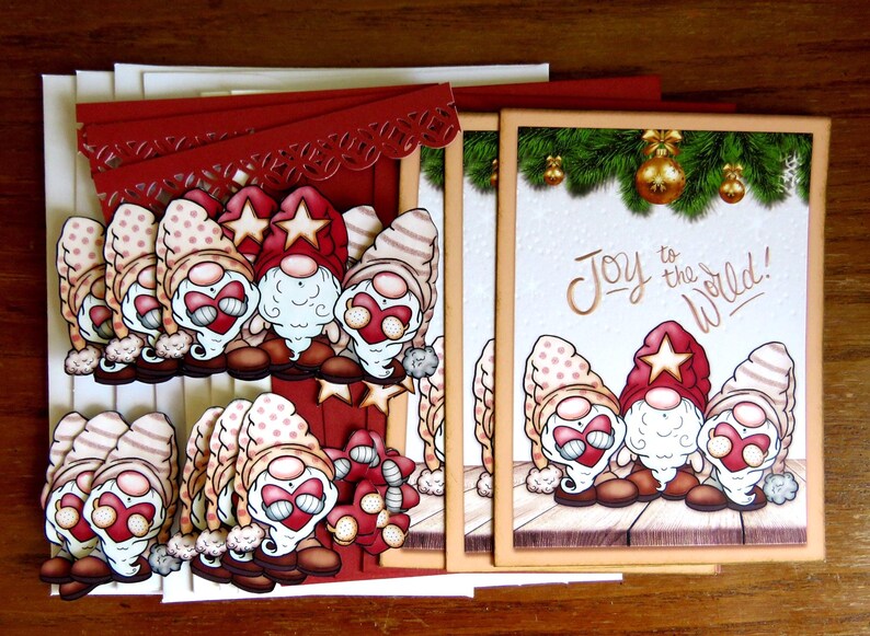 May include: Christmas greeting cards featuring gnome illustrations. The gnomes wear red hats with stars and have white beards. One card has the text "Joy to the World!" and a green border with gold ornaments. The cards are on a wooden surface.