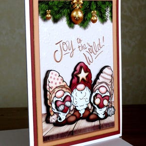 May include: A Christmas greeting card featuring three cartoon gnomes with heart-shaped faces and festive hats. The card has a textured white background with the words "Joy to the World!" and is framed with red and tan borders. Gold ornaments and greenery are at the top.