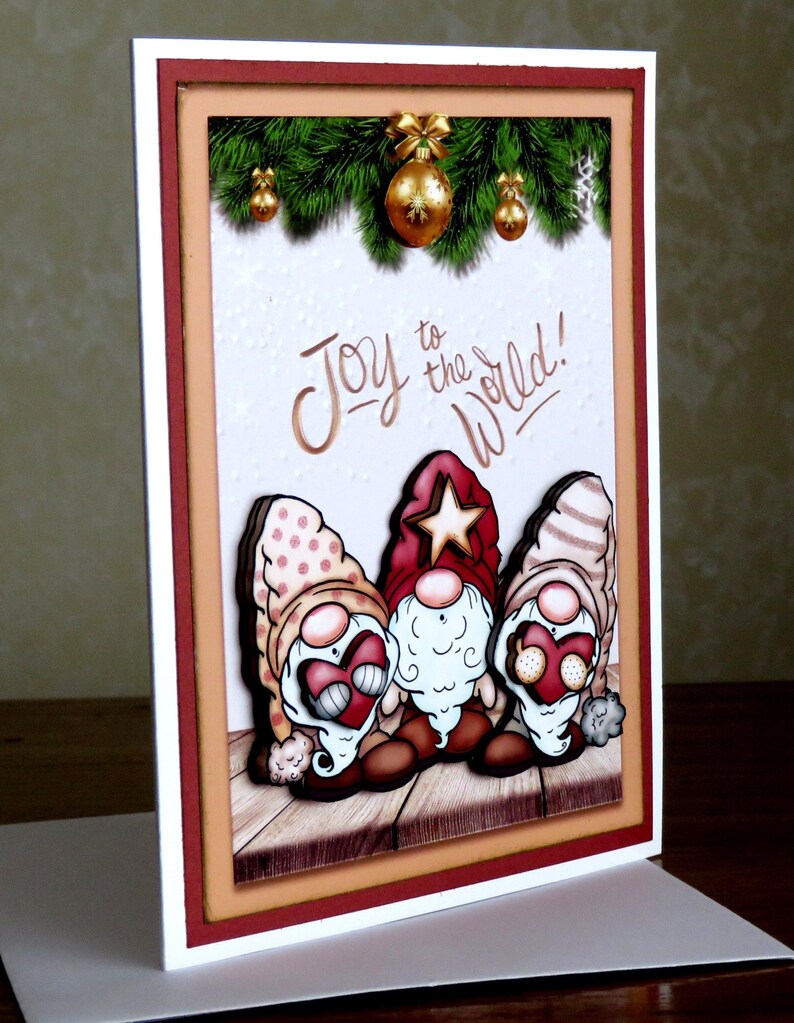 May include: A Christmas greeting card featuring three cartoon gnomes with heart-shaped faces and festive hats. The card has a textured white background with the words "Joy to the World!" and is framed with red and tan borders. Gold ornaments and greenery are at the top.