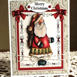 May include: A rectangular Christmas greeting card featuring a central illustration of Santa Claus with a white beard, red coat, and a sack, accented by two red satin bows, a twine bow, and a white label with the text Merry Christmas.