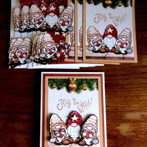 May include: Greeting cards featuring whimsical gnomes with red hats and star accents. The cards have the phrase "Joy to the World!" and are decorated with festive elements like pine boughs and gold ornaments. The cards are on a wooden surface.