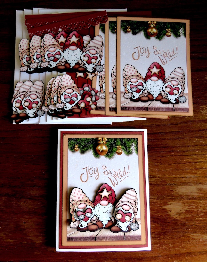 May include: Greeting cards featuring whimsical gnomes with red hats and star accents. The cards have the phrase "Joy to the World!" and are decorated with festive elements like pine boughs and gold ornaments. The cards are on a wooden surface.