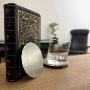 Pode incluir: Um livro preto encadernado em couro com letras douradas em relevo e um design decorativo. Uma base de metal escovado encosta no livro. Um copo de água está sobre uma pilha de bases.