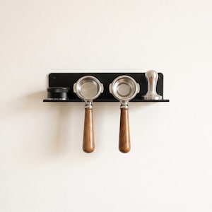 Peut inclure: Un support mural en métal noir contenant deux porte-filtres à expresso avec des poignées en bois, un distributeur de café circulaire noir et un tasseur à café en métal argenté. Le support est rectangulaire avec une étagère plate.