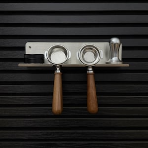 Könnte beinhalten: Ein Wandregal aus Edelstahl mit zwei Espresso-Siebträgern mit braunen Holzgriffen, einem kleinen schwarzen runden Behälter und einem silbernen Kaffeestampfer aus Metall. Das Regal besteht aus einer flachen Ablage mit Rückwand.