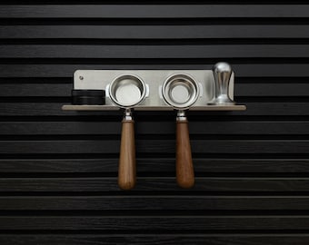 Pode incluir: Um suporte de parede em aço inoxidável contendo dois porta-filtros de café expresso com cabos de madeira castanha, um pequeno recipiente circular preto e um calcador de café em metal prateado. O suporte é uma prateleira horizontal.