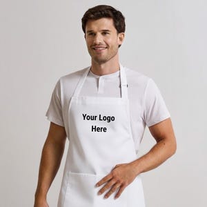 Puede incluir: Un delantal blanco liso con una correa para el cuello y un bolsillo frontal, con el texto negro "Your Logo Here" impreso en el centro del área del pecho. El delantal está confeccionado con una tela blanca sólida e incluye un lazo.