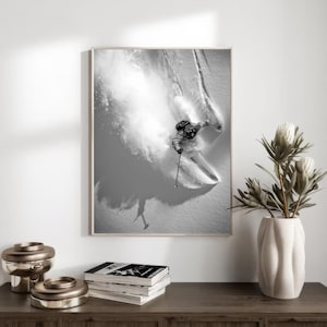 May include: A black and white photograph of a skier carving through fresh powder, creating a plume of snow behind them.