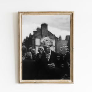 May include: A black and white photograph of a man blowing bubbles in front of a building. The man is wearing a suit and tie, and he is looking up at the bubbles. The building is a brick building with a few windows.