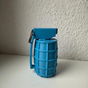Könnte beinhalten: Eine blaue Spielzeuggranate mit Griff und Ring. Die Granate hat einen zylindrischen Körper mit gitterartigem Muster und eine rechteckige Oberseite. Griff und Ring sind metallisch.