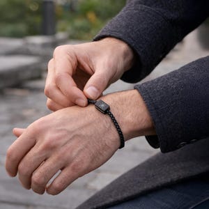 May include: A black bracelet with a square centerpiece featuring a sunburst design. The bracelet is made of a chain link style and is being fastened on a wrist. The person is wearing a dark gray coat.